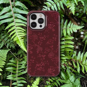 May include: A burgundy phone case with a floral pattern. The case features a detailed design of roses and other flowers. The phone case is surrounded by green ferns.