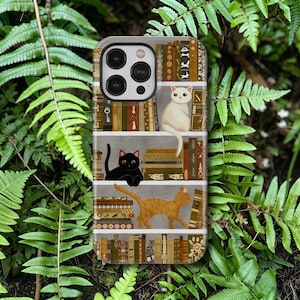 May include: A smartphone case featuring a graphic illustration of three cats on bookshelves filled with various books. A white cat sits on the top shelf, a black cat rests on the middle shelf, and an orange tabby cat walks on the lower shelf.