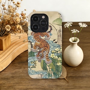 May include: A phone case featuring a detailed illustration of a tiger with orange and black stripes, set against a backdrop of water, flowers, and Japanese-style art. The case has a camera cutout and a smooth finish.