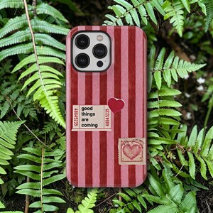 Puede incluir: Funda de teléfono con un diseño de rayas verticales rojas y rosas. La funda presenta un pequeño corazón rojo, un ticket que dice "good things are coming" y un sello con un diseño de corazón.