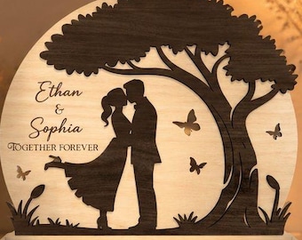 May include: A semi-circular wooden light display featuring a dark brown laser-cut silhouette of a couple standing under a tree. The light-colored background includes the text Ethan & Sophia and TOGETHER FOREVER, with small butterfly cutouts.