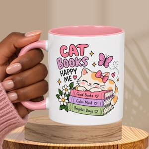 May include: A white ceramic mug with a pink interior and handle, featuring a graphic of a cat resting on a stack of three books labeled Good Books, Calm Mind, and Brighter Days, with the text CAT BOOKS HAPPY ME, flowers, and a butterfly.