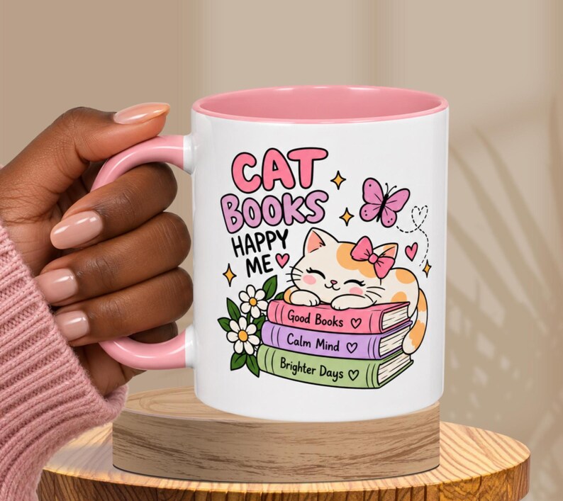 May include: A white ceramic mug with a pink interior and handle, featuring a graphic of a cat resting on a stack of three books labeled Good Books, Calm Mind, and Brighter Days, with the text CAT BOOKS HAPPY ME, flowers, and a butterfly.