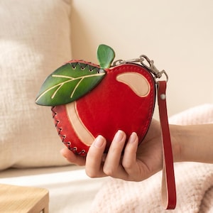 May include: A red leather apple-shaped coin purse with a green leaf and a wrist strap. The purse has a zipper closure and a light-colored accent on the side. The leaf is detailed with green and yellow veins.
