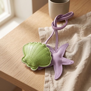 May include: A light green seashell-shaped leather keychain and a lavender starfish-shaped leather keychain are connected by a lavender leather loop. The keychains are on a wooden surface next to a beige cloth.