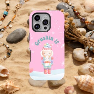 Puede incluir: Funda de teléfono rosa con un diseño de cascanueces de dibujos animados y el texto "Crushin it". El cascanueces lleva un atuendo azul y rosa, un sombrero y sostiene un postre en forma de cono. Los copos de nieve están esparcidos por el diseño.