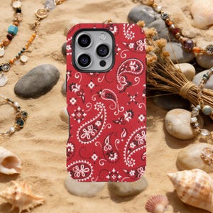 May include: A red phone case with a paisley pattern in white and black. The case is on a sandy surface with jewelry and seashells in the background. The case has a camera module with three lenses.