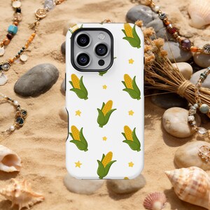 Puede incluir: Una funda de teléfono blanca con un patrón repetido de mazorcas de maíz amarillas con hojas verdes y formas de estrella amarillas. La funda del teléfono tiene un módulo de cámara negro y está sobre arena.