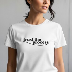 Puede incluir: Camiseta blanca con la frase "trust the process" en negro, arqueada sobre una línea curva. La camiseta es de manga corta y está hecha de un material suave. El tema general es motivacional.