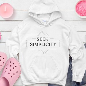 Puede incluir: Sudadera con capucha blanca con el texto "SEEK SIMPLICITY" en un diseño geométrico. La imagen incluye zuecos rosas, una vela, un cuenco de gránulos rosas, pantalones cortos vaqueros y rosas rosas, todo dispuesto sobre una superficie de madera blanca.