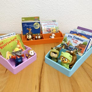 Puede incluir: Tres organizadores de libros coloridos llenos de libros infantiles y figuritas. Los organizadores son de color morado, naranja y azul. Los libros tienen varios títulos e ilustraciones, y las figuritas representan animales y personajes de dibujos animados.