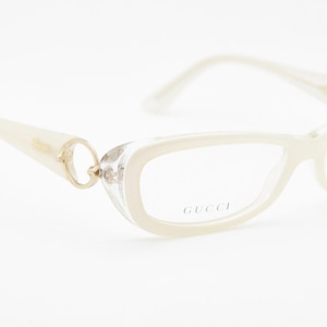 Puede incluir: Gafas Gucci de color marfil con forma de ojo de gato. Las monturas tienen un aspecto translúcido con detalles dorados en las patillas y bisagras. El logotipo de Gucci es visible en la lente. Las gafas están sobre un fondo blanco.