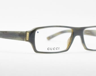 Puede incluir: Gafas de montura rectangular con un patrón veteado gris y verde oliva. El logotipo de Gucci es visible en la montura. Las gafas tienen lentes transparentes y un acento dorado en las patillas.