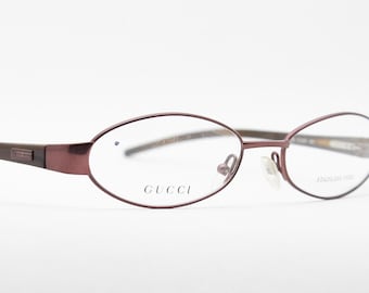 Puede incluir: Gafas ovaladas con montura burdeos y varillas marrones oscuras. El nombre de la marca "GUCCI" está impreso en la lente. Las gafas tienen un diseño minimalista y están sobre un fondo blanco.