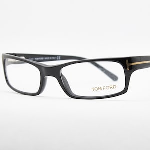 Puede incluir: Gafas rectangulares negras con lentes transparentes. Las monturas tienen un acento dorado en las patillas y la marca "TOM FORD" está impresa en la lente. Las gafas están sobre un fondo blanco.