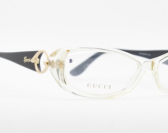 Puede incluir: Gafas con montura transparente, varillas negras y detalles dorados. Las gafas tienen forma de ojo de gato y la marca "GUCCI" está impresa en la lente.