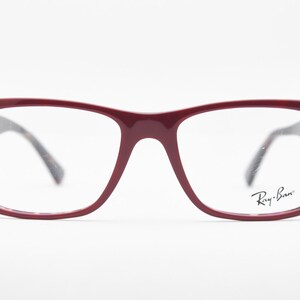 May include: Burgundy-colored Ray-Ban eyeglasses with a rectangular frame. The glasses have clear lenses and a subtle Ray-Ban logo on the right lens. The temples are a dark tortoiseshell pattern. The glasses are set against a white background.