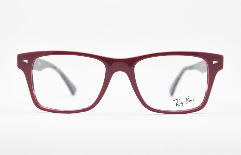 May include: Burgundy-colored Ray-Ban eyeglasses with a rectangular frame. The glasses have clear lenses and a subtle Ray-Ban logo on the right lens. The temples are a dark tortoiseshell pattern. The glasses are set against a white background.