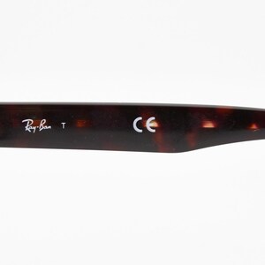 May include: Close-up of a Ray-Ban eyeglass arm. The arm is a dark brown tortoiseshell pattern with the Ray-Ban logo and the letters "CE" printed on it. The arm has a silver metal hinge.