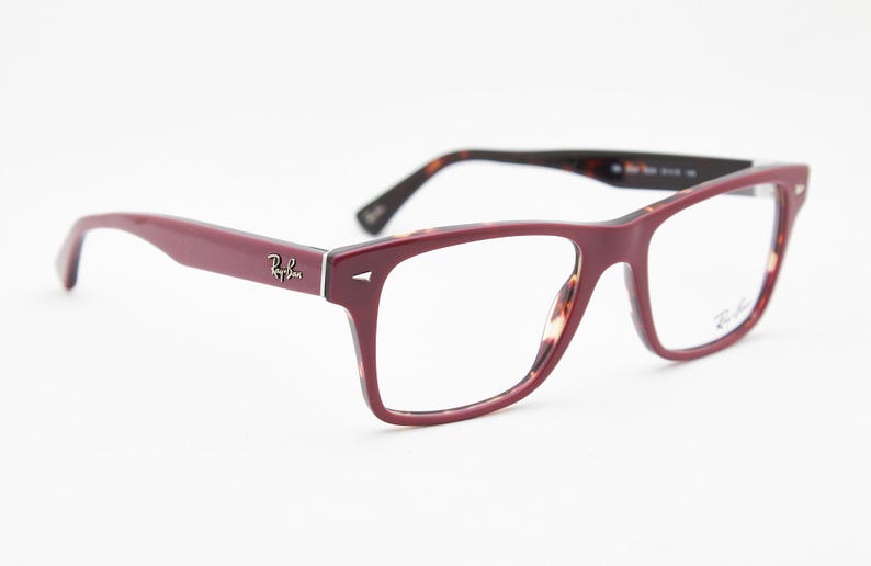 May include: Burgundy-colored Ray-Ban eyeglasses with a rectangular frame. The temples are a solid burgundy, while the inner frame has a tortoiseshell pattern. The Ray-Ban logo is visible on the temple and lens.
