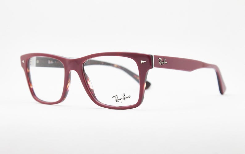 May include: Burgundy-colored Ray-Ban eyeglasses with a rectangular frame. The temples and frame are a solid burgundy color, with the Ray-Ban logo on the temple and lens. The interior of the frame has a tortoiseshell pattern.