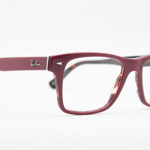 May include: Burgundy-colored Ray-Ban eyeglasses with a rectangular frame. The temples and frame are a solid burgundy color, with the Ray-Ban logo on the temple. The interior of the frame has a tortoiseshell pattern.