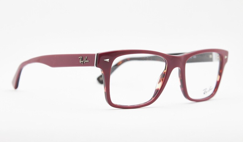 May include: Burgundy-colored Ray-Ban eyeglasses with a rectangular frame. The temples and frame are a solid burgundy color, with the Ray-Ban logo on the temple. The interior of the frame has a tortoiseshell pattern.