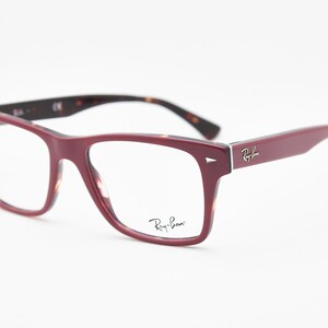 May include: Burgundy-colored eyeglasses with a rectangular frame. The temples and inner frame feature a tortoiseshell pattern. The Ray-Ban logo is visible on the temple and lens. The glasses are set against a white background.