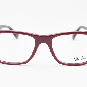 May include: Burgundy-colored eyeglasses with a rectangular frame. The glasses have a tortoiseshell pattern on the inner frame and temples. The Ray-Ban logo is visible on the right lens.