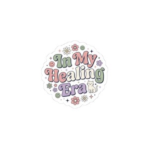 May include: A round sticker with the text "In My Healing Era" in a retro font. The sticker features a white cat, flowers, and smiley faces in pastel colors, including pink, green, purple, and orange. The sticker has a white border.