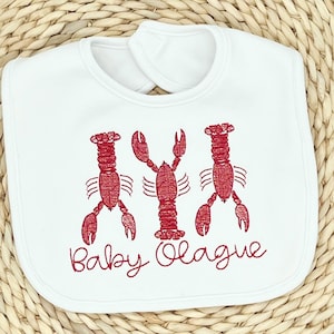 May include: White baby bib with three red embroidered lobsters and the text "Baby O'Lague" in red cursive. The bib has a rounded shape with a neck opening for easy wear. The design is simple and playful.