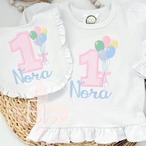 May include: White long-sleeved shirt and bib set with a pink striped number 1 and the name "Nora" in light blue. The shirt and bib feature a ruffle trim and a design of colorful balloons.