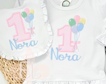 May include: White long-sleeved shirt and bib set with a pink striped number 1 and the name "Nora" in light blue. The shirt and bib feature a ruffle trim and a design of colorful balloons.