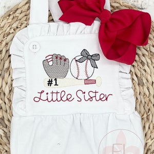 May include: White baby onesie with a red bow, embroidered with a baseball, bat, glove, and the text "#1 Little Sister".