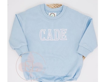 May include: A pale blue baby romper featuring the name "CADE" embroidered in white. The romper has long sleeves and a round neckline. The bottom has snap closures.