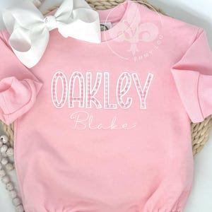 May include: A pink baby bubble romper with long sleeves and a white bow. The name "Oakley" is embroidered in white gingham letters, with the name "Blake" embroidered below in cursive. The romper is on a woven mat.