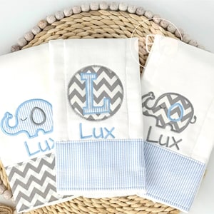 May include: Three white baby burp cloths with blue and gray elephant and chevron designs. Each cloth has a blue striped bottom and the name "Lux" embroidered in blue. One cloth features the letter "L".
