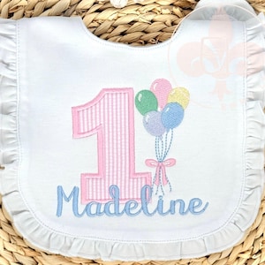 May include: White baby bib with a ruffled edge, featuring a pink striped number 1 and a cluster of colorful balloons. The name "Madeline" is embroidered in light blue script, making it a personalized birthday accessory.