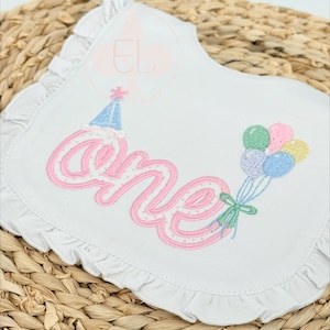 May include: A white fabric bib with a ruffled edge featuring embroidered text that reads one in pink. A blue party hat sits atop the letter o, and a cluster of pastel-colored balloons in green, pink, yellow, and blue is tied with a green ribbon.