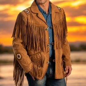 May include: A tan suede jacket featuring long fringe detailing across the chest, shoulders, and sleeves. The jacket includes silver-toned buttons, decorative beaded patches on the shoulders and elbows, and a blue denim shirt worn underneath.