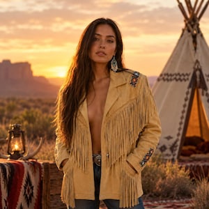 May include: A tan suede jacket featuring long fringe detailing across the chest and sleeves, a notched lapel, and colorful geometric embroidery on the shoulders and cuffs. The jacket is worn open over blue denim jeans with a silver belt buckle.