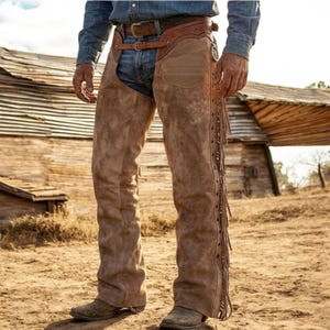 Puede incluir: Chaps de cuero de ante color canela con un canesú de cuero labrado en la cintura y cierre de hebilla. Los chaps tienen flecos largos a lo largo de las piernas exteriores, detalles de tachuelas metálicas y un acabado liso, sobre vaqueros azules.