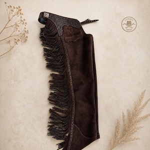 Peut inclure: Un chaps en cuir suédé marron foncé avec de longues franges sur le côté, une sangle à boucle réglable sur le haut et un motif floral gaufré sur l'empiècement supérieur. Un petit logo circulaire avec le texte COWBOY CREST est visible.