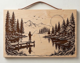 May include: Wooden wall art featuring a detailed engraving of a serene lake scene. A person is fishing from a dock, surrounded by trees, mountains, and a full moon. The artwork includes a brown leather strap for hanging.
