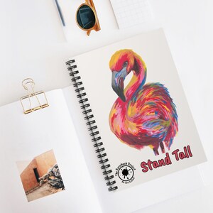 May include: Spiral-bound notebook featuring a vibrant flamingo illustration in shades of red, yellow, and blue. The words "Stand Tall" are printed in pink. The notebook is on a white surface with a gold binder clip and sunglasses.