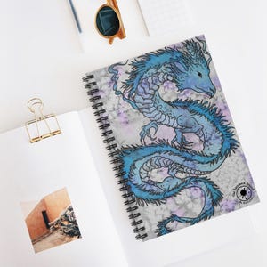 May include: Spiral-bound notebook featuring a blue dragon illustration on a tie-dye background. The dragon is detailed with shades of blue and black. The notebook has a black spiral binding and the text "The Kotteck Project" and "An Art Collab" on the bottom right corner.
