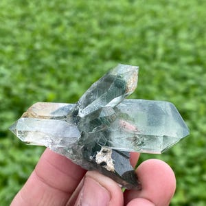 May include: A cluster of clear quartz crystals with green and brown inclusions. The crystals are held in a hand, with a blurred green background. The quartz has a natural, rough texture and a variety of shapes, with sharp points and facets.