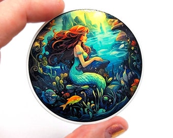 May include: A round sticker with a colorful illustration of a mermaid sitting on a rock in the ocean. The mermaid has long red hair and a green tail. The ocean is blue and green, and there are coral reefs and fish in the background.