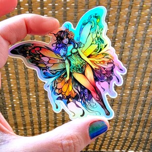 May include: A holographic sticker featuring a fairy with iridescent wings, a green bodice, and a flowing blue hair. The fairy is standing on a flower with a yellow and orange butterfly on her wings.