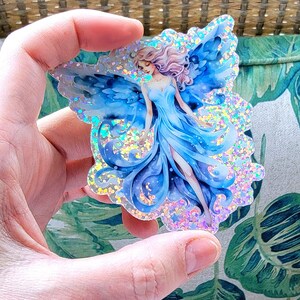 May include: A blue and silver glitter fairy sticker with iridescent wings and a flowing dress. The sticker is on a clear background.